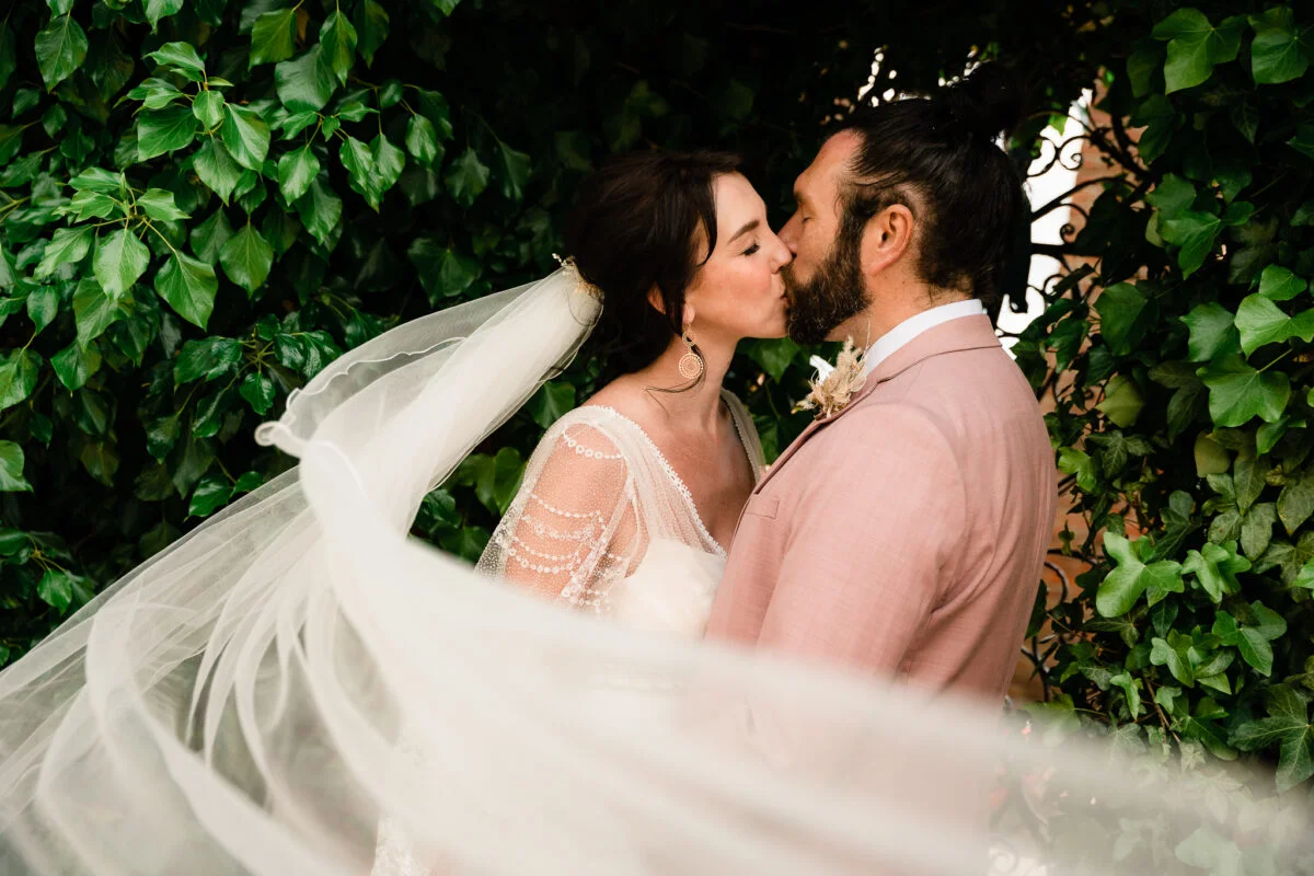 A bride and groom kiss in front of an ivy covered wall.