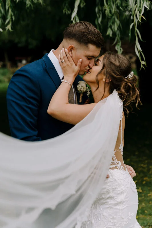 A bride and groom kiss under a willow tree.