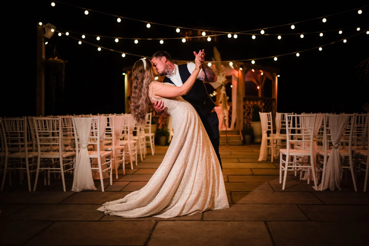 A bride and groom kissing under string lights at their wedding.