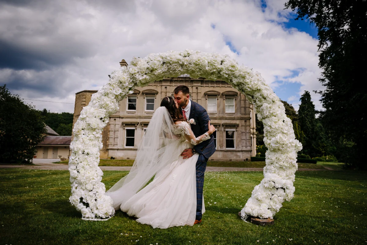 A bride and groom kiss in front of a wedding arch.
