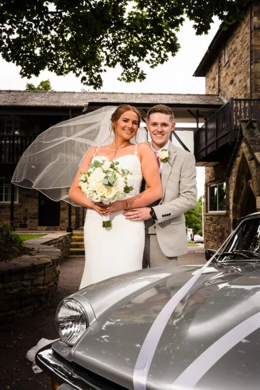 A bride and groom standing next to a silver sports car.