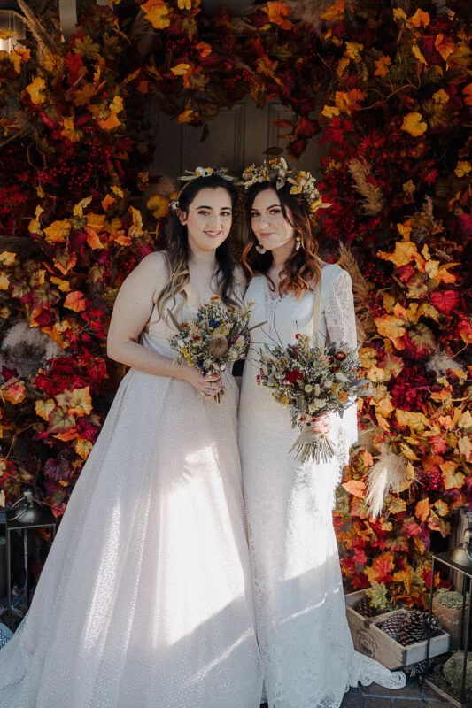 Two brides standing in front of a door decorated with fall leaves.