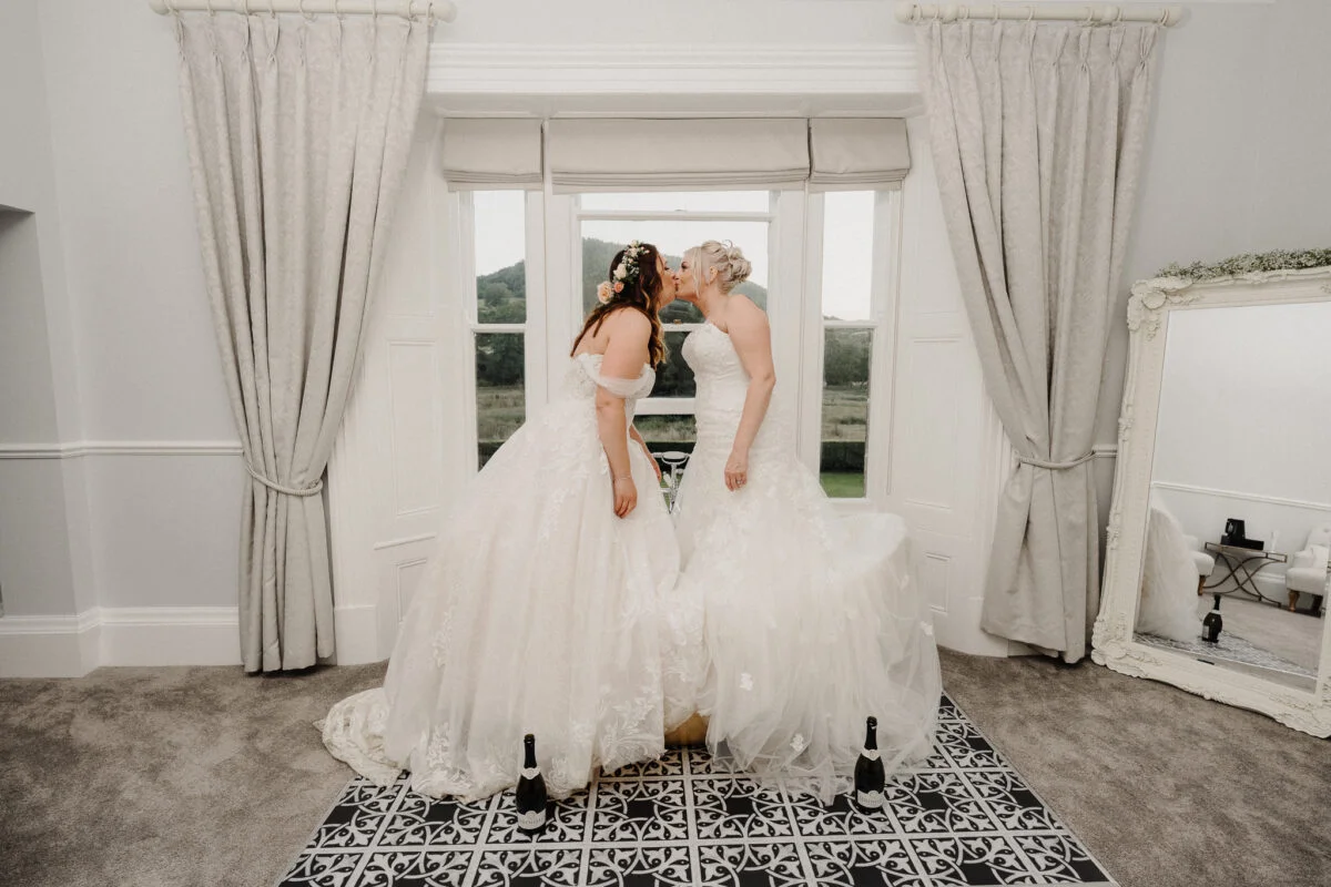 Two brides kissing in front of a mirror in a room.