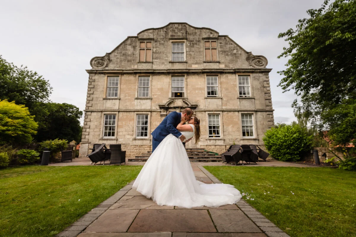 A bride and groom kissing in front of a large mansion.