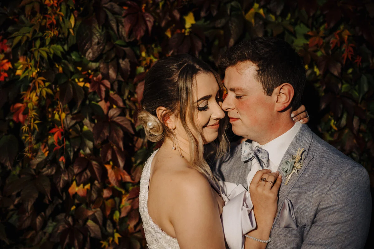 A bride and groom embracing in front of a wall of autumn leaves.
