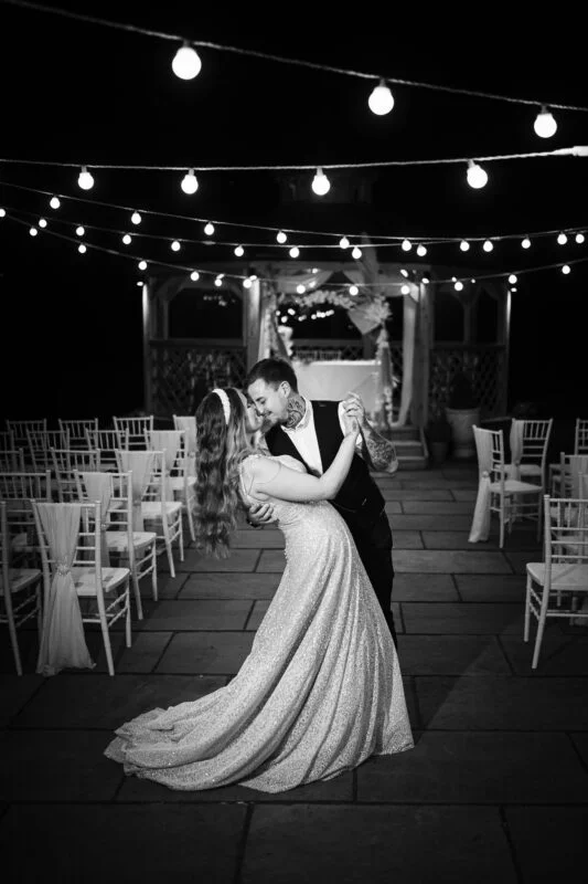Black and white photo of bride and groom kissing under string lights.