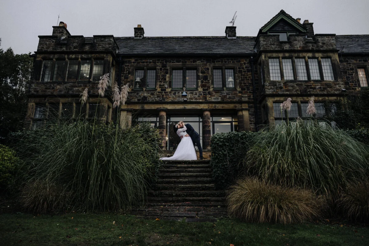 A bride and groom standing in front of a large mansion.