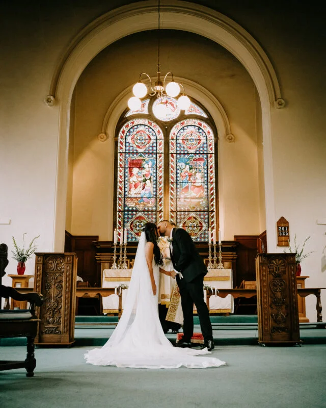 A bride and groom kissing in front of a stained glass window.