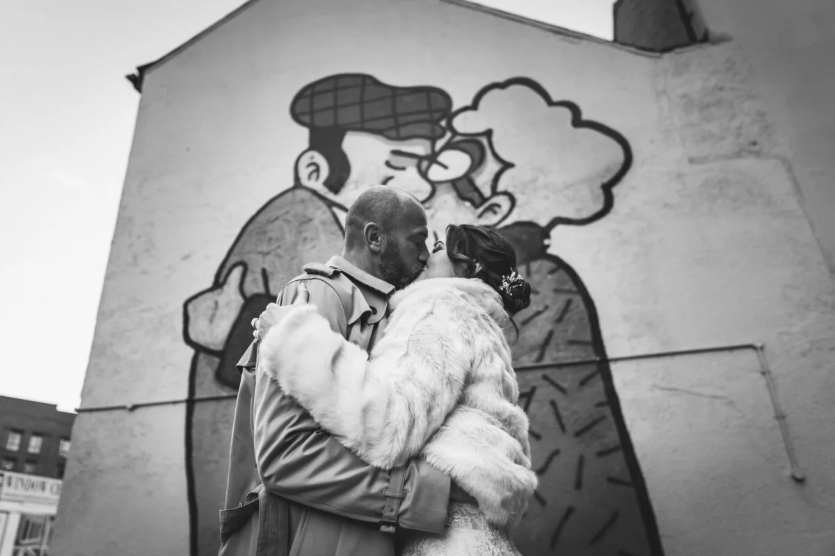 A black and white photo of a couple kissing in front of a mural.