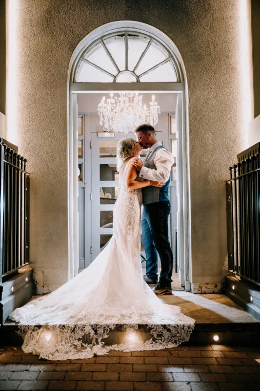 A bride and groom kissing in front of an archway at night.
