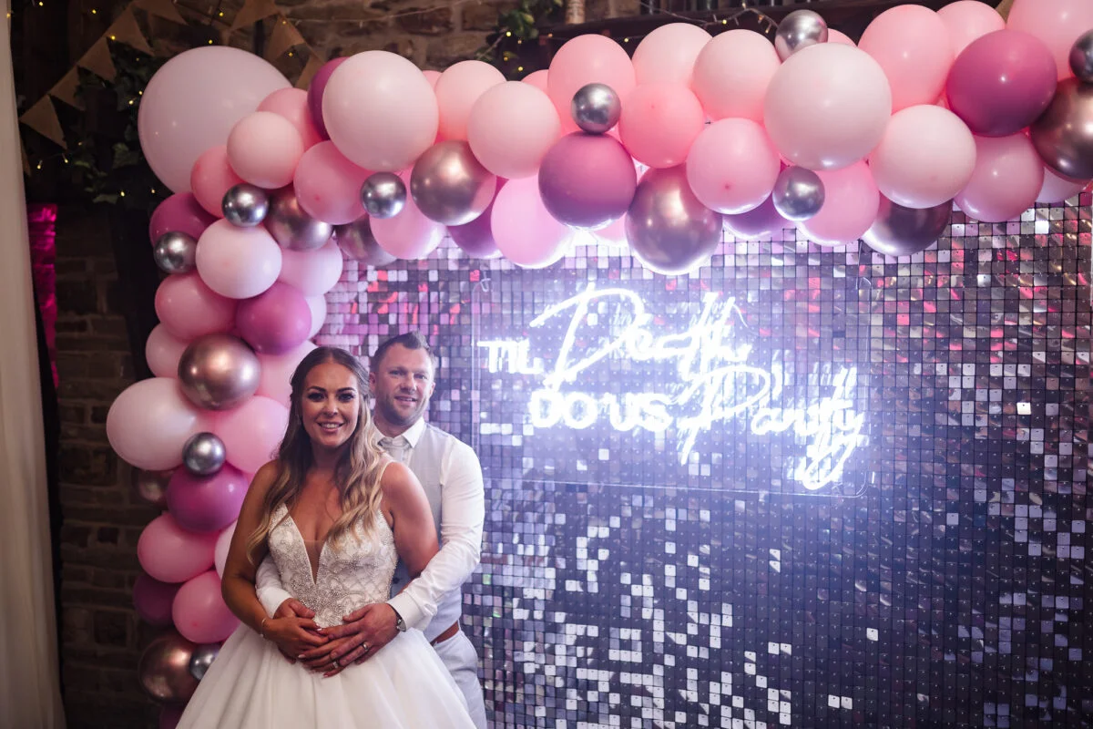 A bride and groom standing in front of a pink balloon arch.