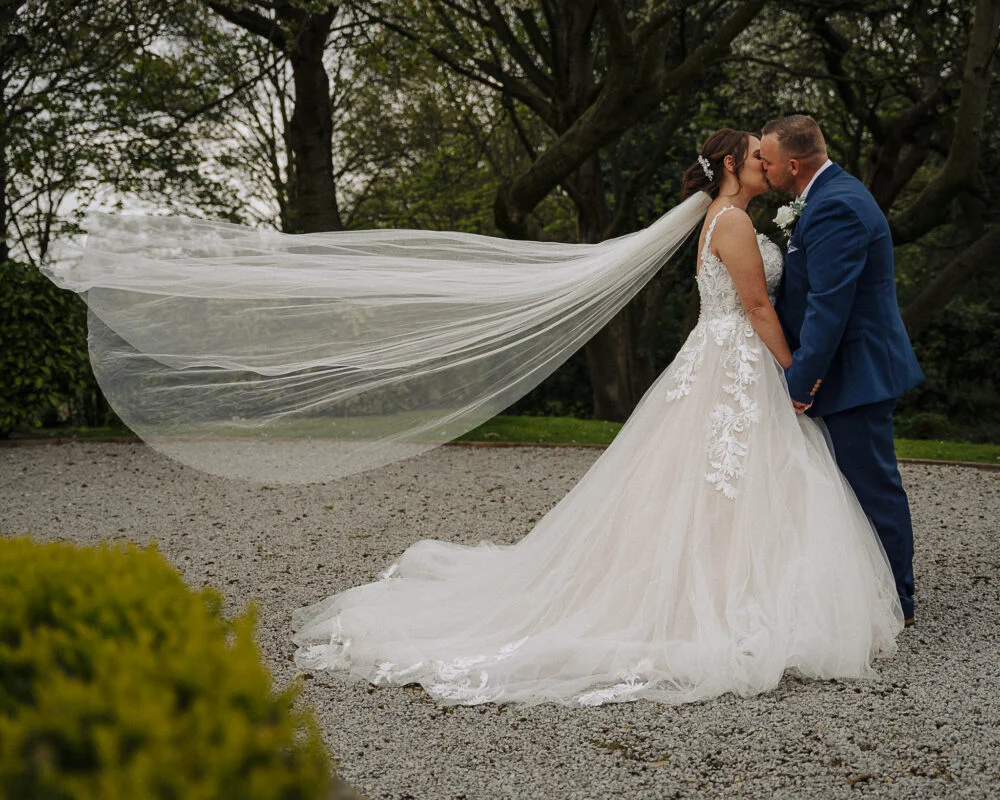 A bride and groom kissing in front of a tree with their veil blowing in the wind.