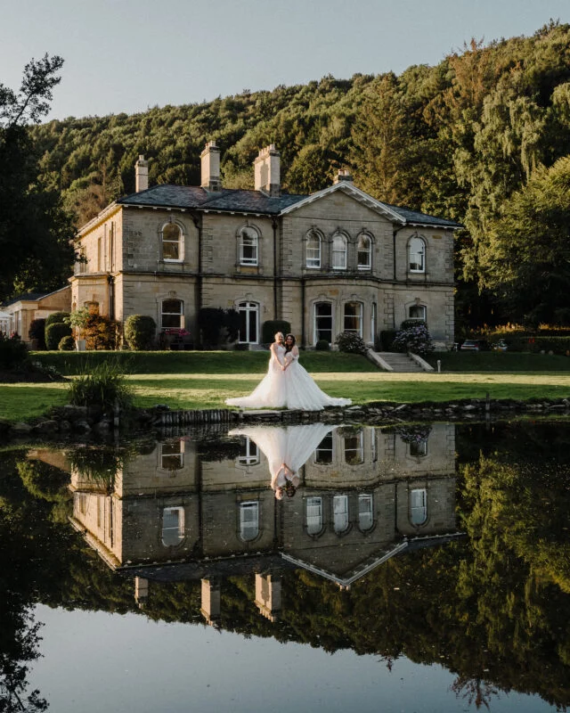 A bride and groom standing in front of a large mansion with a pond in the background.