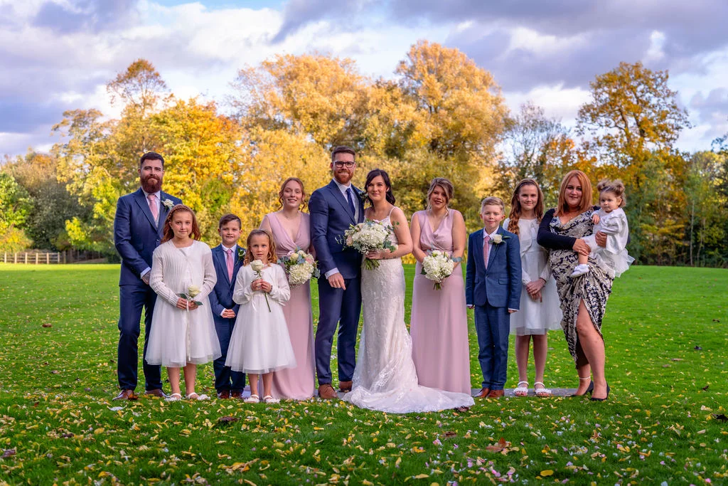 A newlywed couple poses outdoors on a grassy lawn with their large wedding party and family members against a backdrop of golden autumn trees.