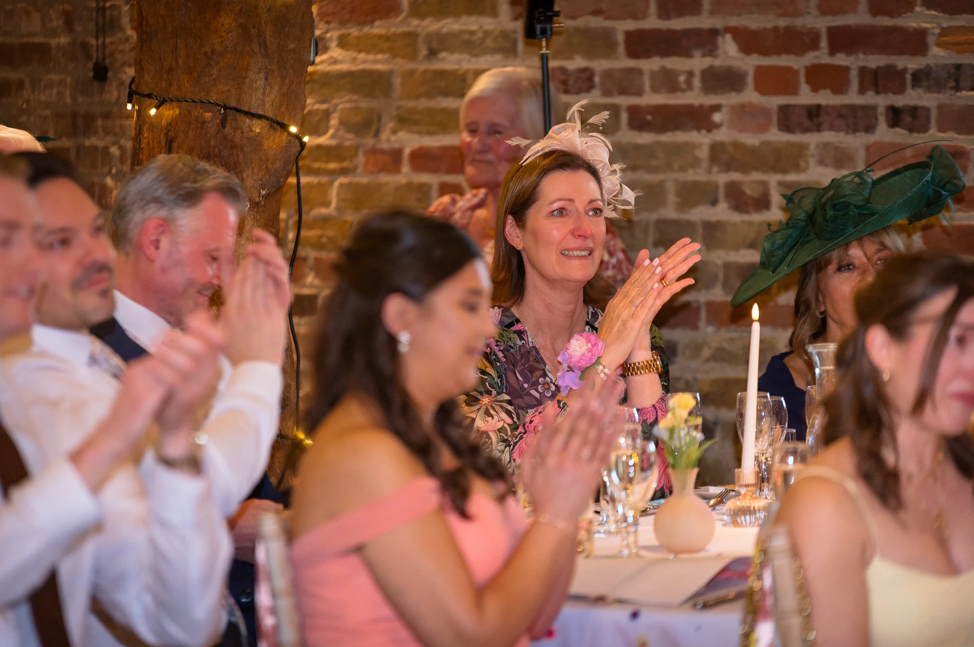 Wedding guests sit at long banquet tables inside a rustic barn with exposed brick walls, clapping and reacting emotionally during speeches.