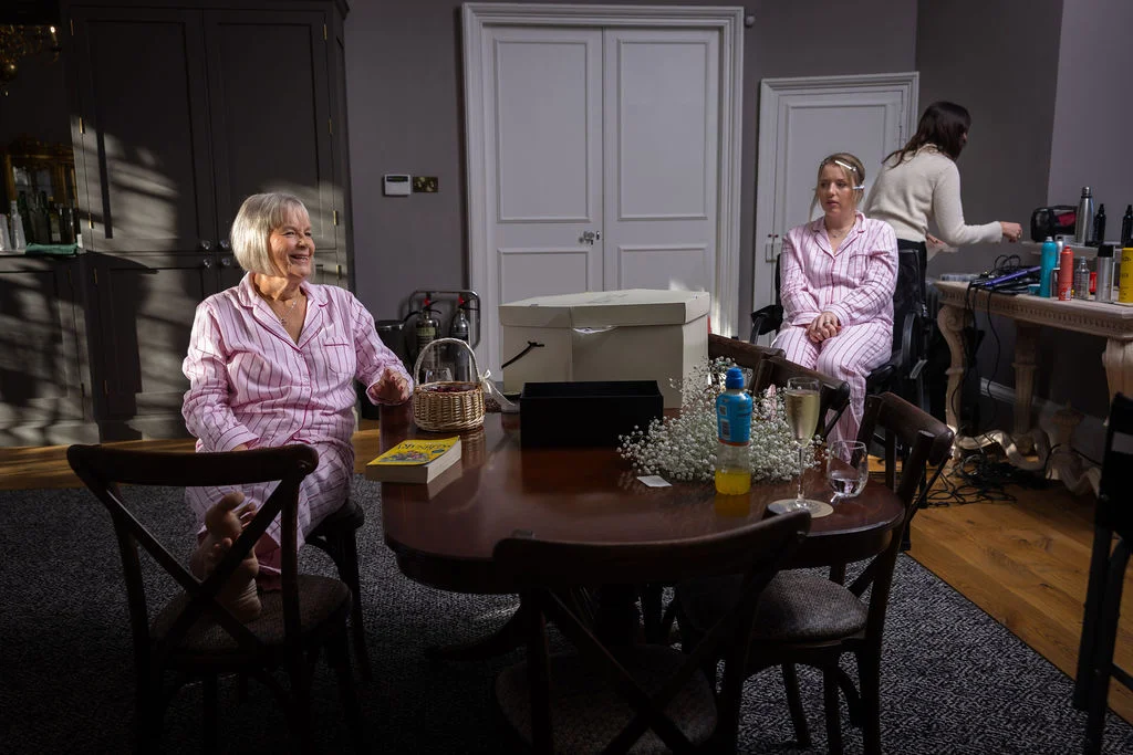 An older woman in matching pink striped pyjamas smiles happily while sitting at a wooden table during bridal morning preparations.