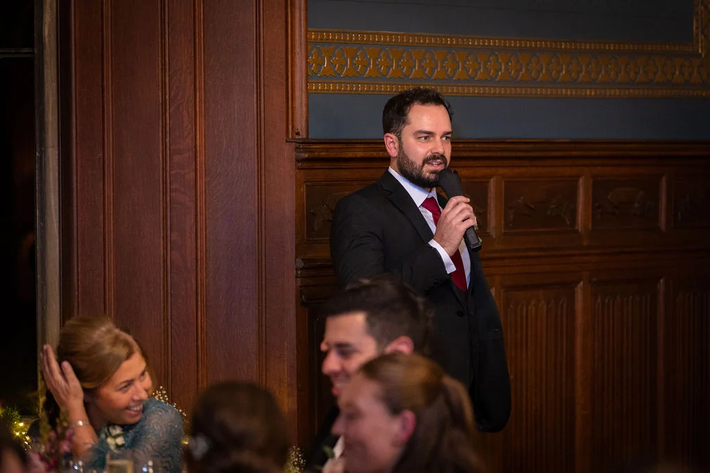 A groom delivers a speech into a microphone in a wood-panelled room while wedding guests sit and listen at the reception.