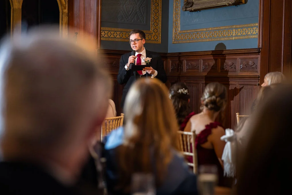 A groom gives a speech holding a microphone and notes in a wood-panelled reception room, viewed over the shoulders of seated wedding guests.