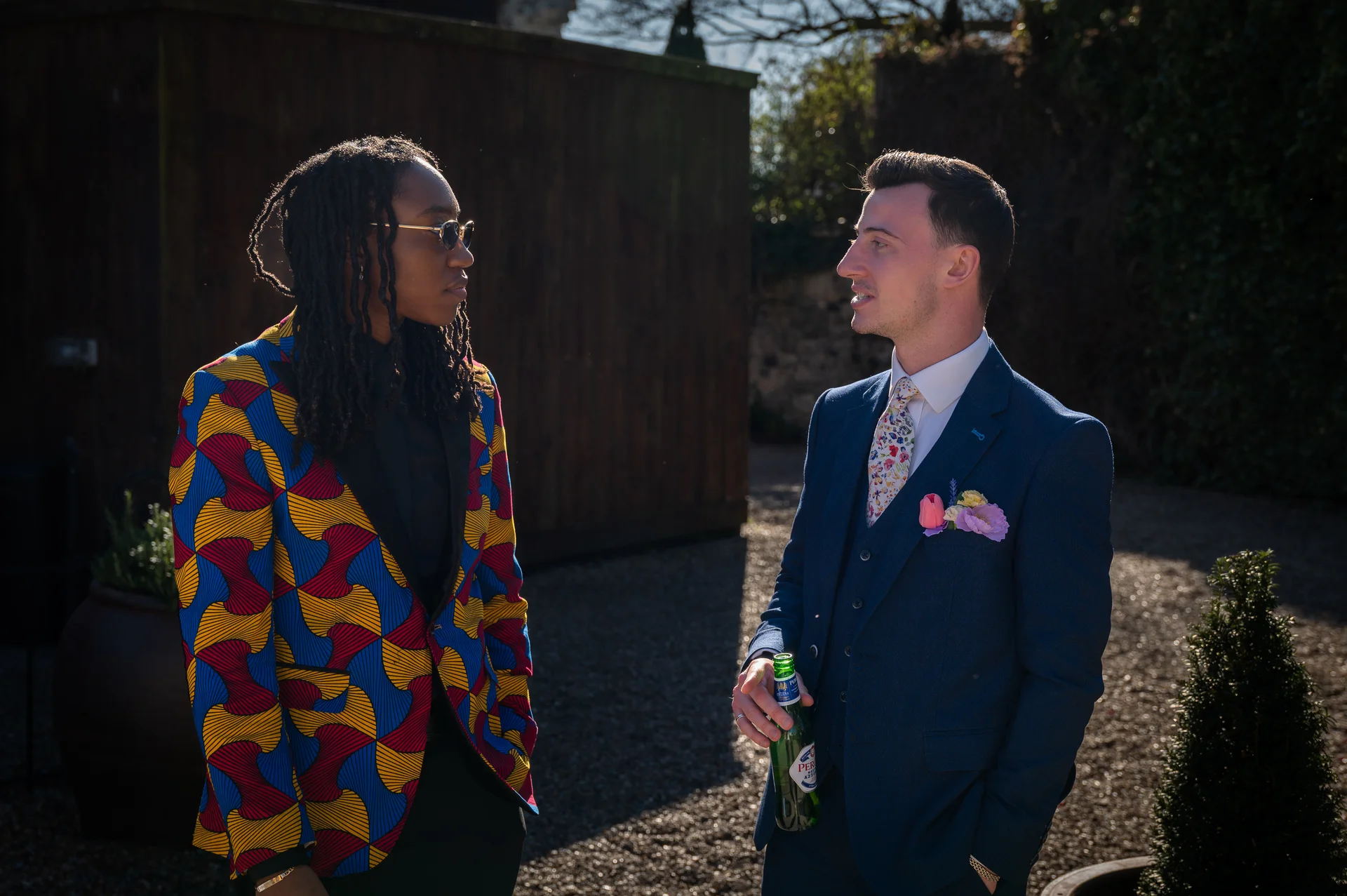 Two grooms stand outside in the bright sunshine, one wearing a vibrant patterned jacket and sunglasses while the other wears a classic navy suit and holds a bottled drink.