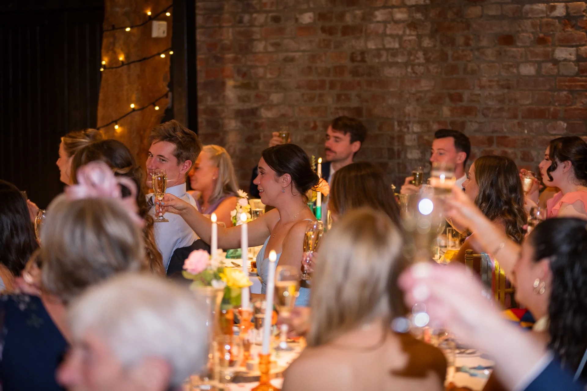 Wedding guests raise their glasses for a toast during the wedding breakfast in a candlelit room featuring exposed brick walls and warm ambient lighting.