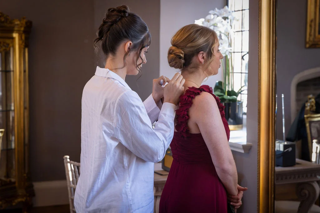 A bridesmaid in a white getting-ready shirt fastens a burgundy bridesmaid dress for another bridesmaid in front of a mirror.
