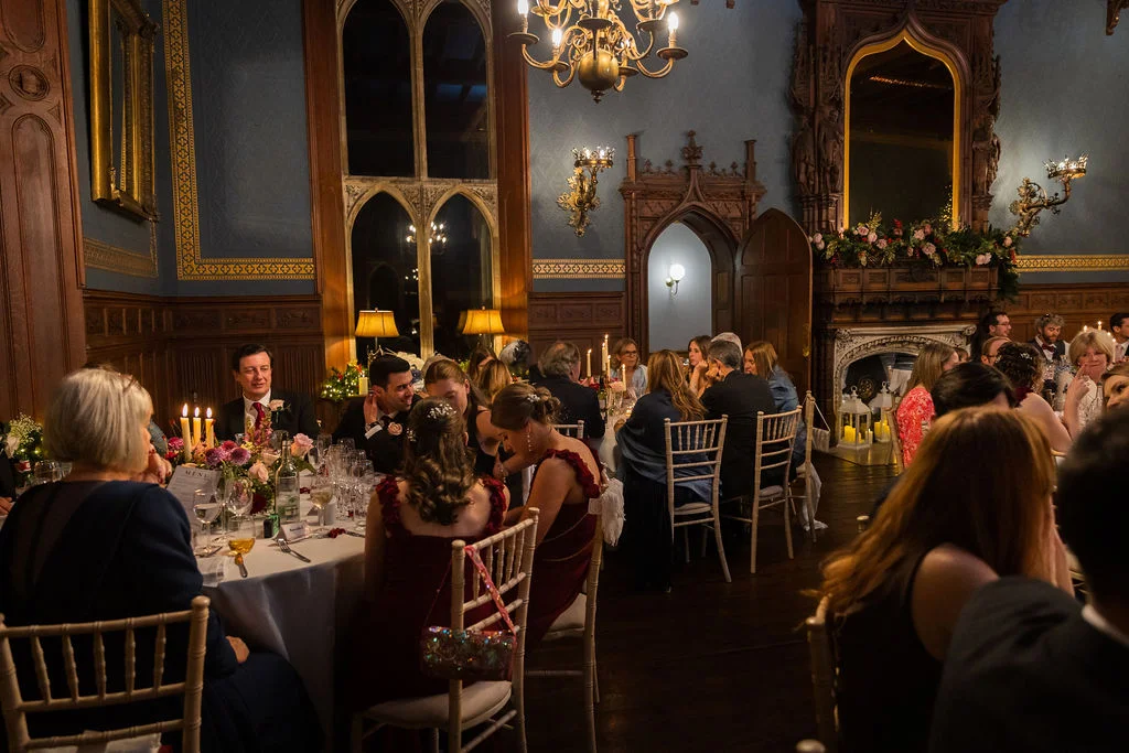 Guests enjoy a warm candlelit wedding breakfast seated at round tables inside a grand stately home featuring wood-panelled walls and tall arched windows.