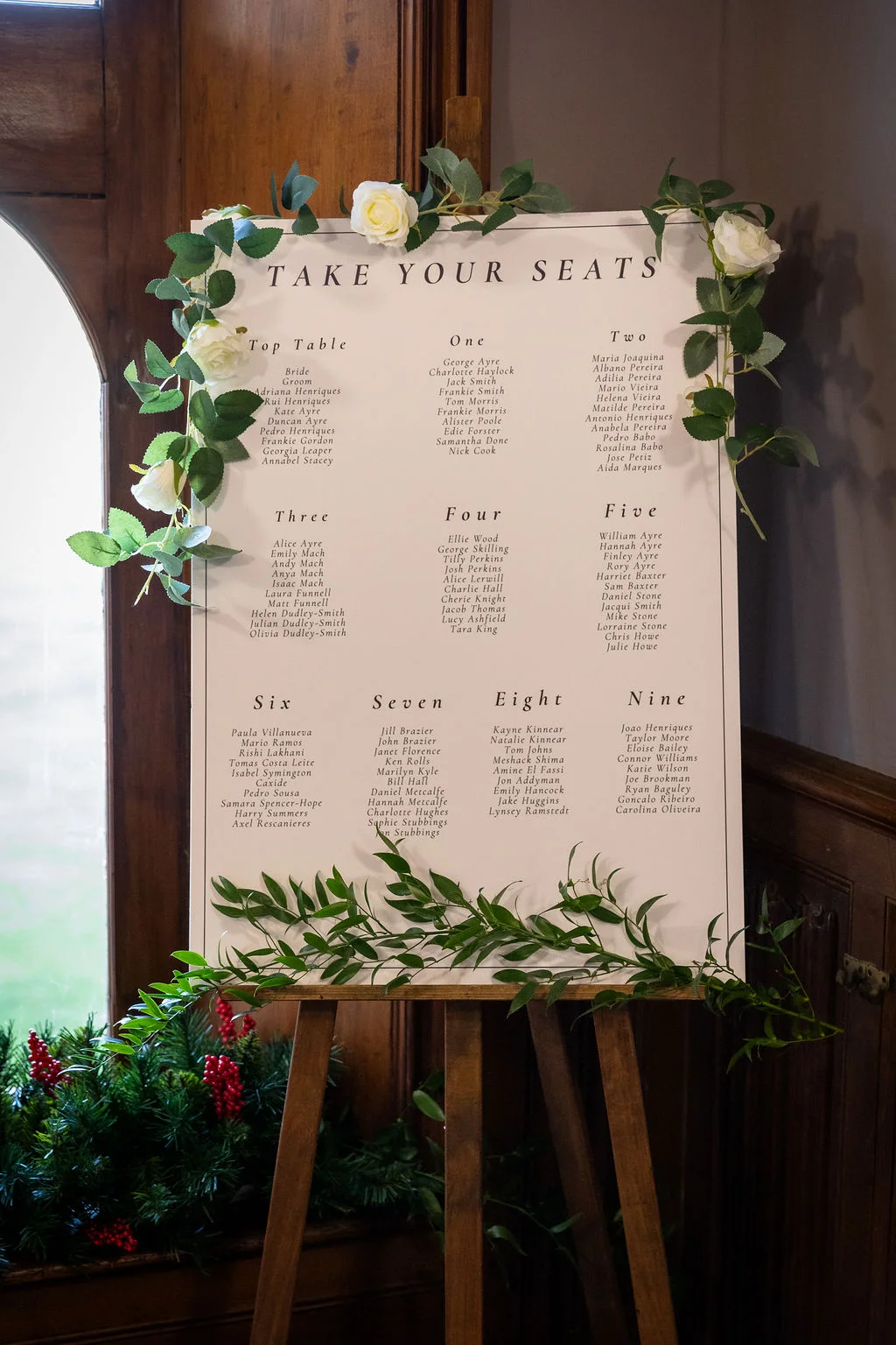 An elegant wedding seating plan board displaying guest names is mounted on a wooden easel and decorated with white roses and trailing eucalyptus leaves.