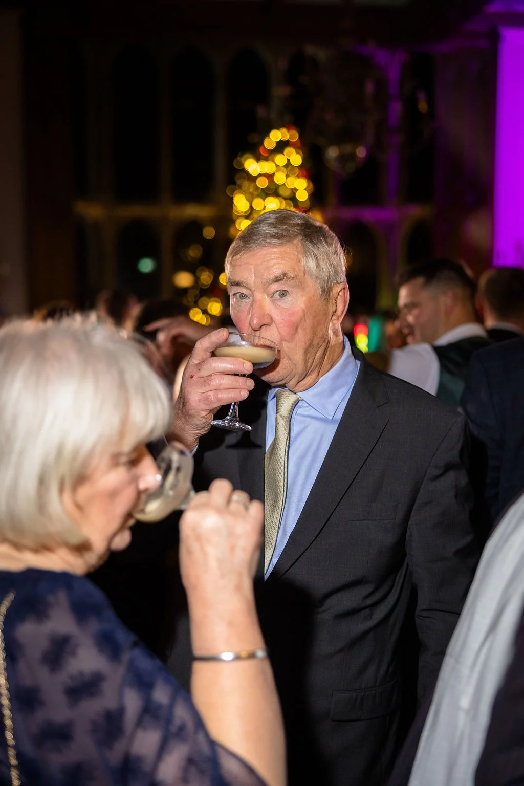 An older male guest in a black suit and patterned tie drinks from a champagne coupe during an evening wedding reception with festive fairy lights.