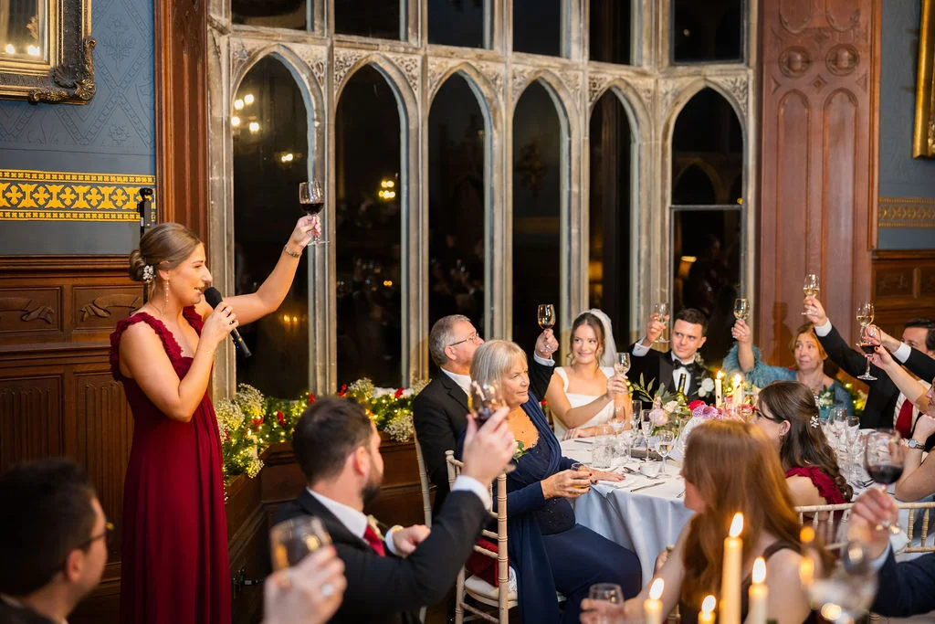 A bridesmaid in a burgundy dress delivers a speech while holding a glass of wine to toast at a candlelit wedding reception in a historic hall.