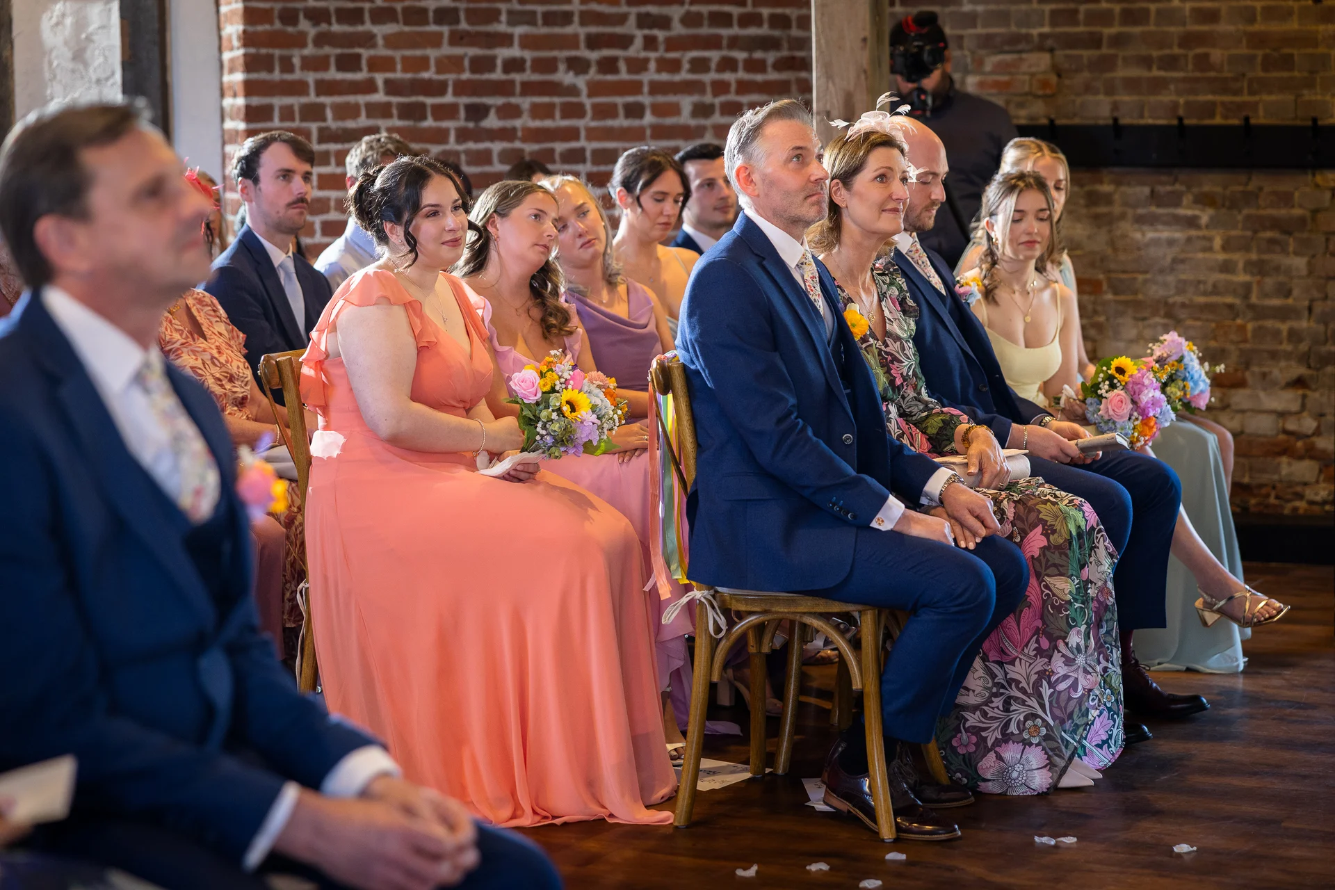 A candid shot of seated wedding guests attentively watching a ceremony inside a venue with exposed brick walls and wooden flooring.