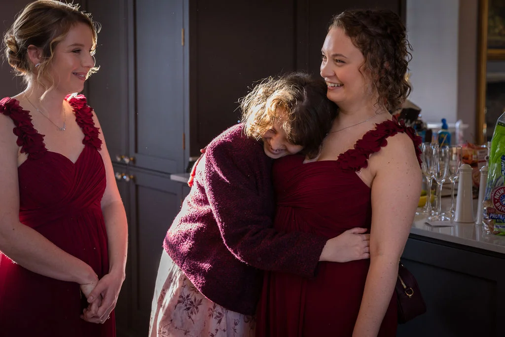A joyful bridesmaid shares a warm hug with another woman indoors while a second bridesmaid in a matching burgundy gown smiles on.