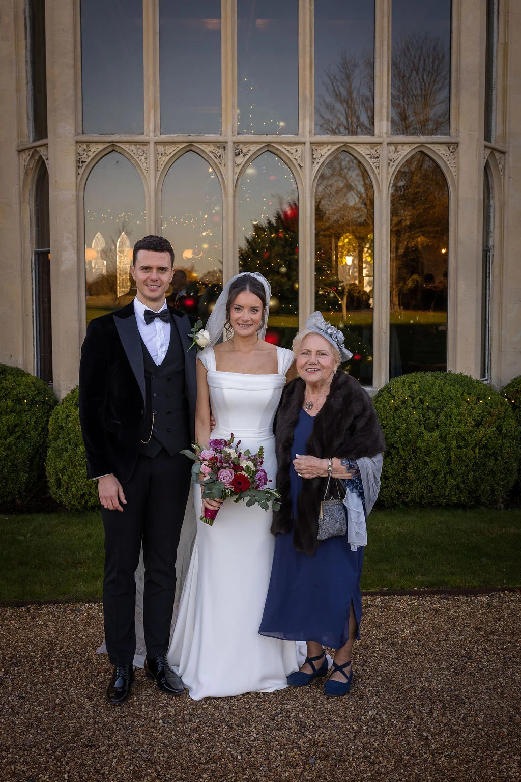 A newlywed couple poses outside a historic manor house with an elderly female relative during a winter wedding, with the bride holding a colourful bouquet.