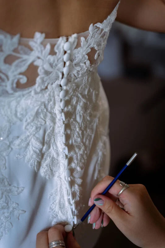 Close-up of a bride fastening the delicate covered buttons down the intricate lace illusion back of her wedding gown.
