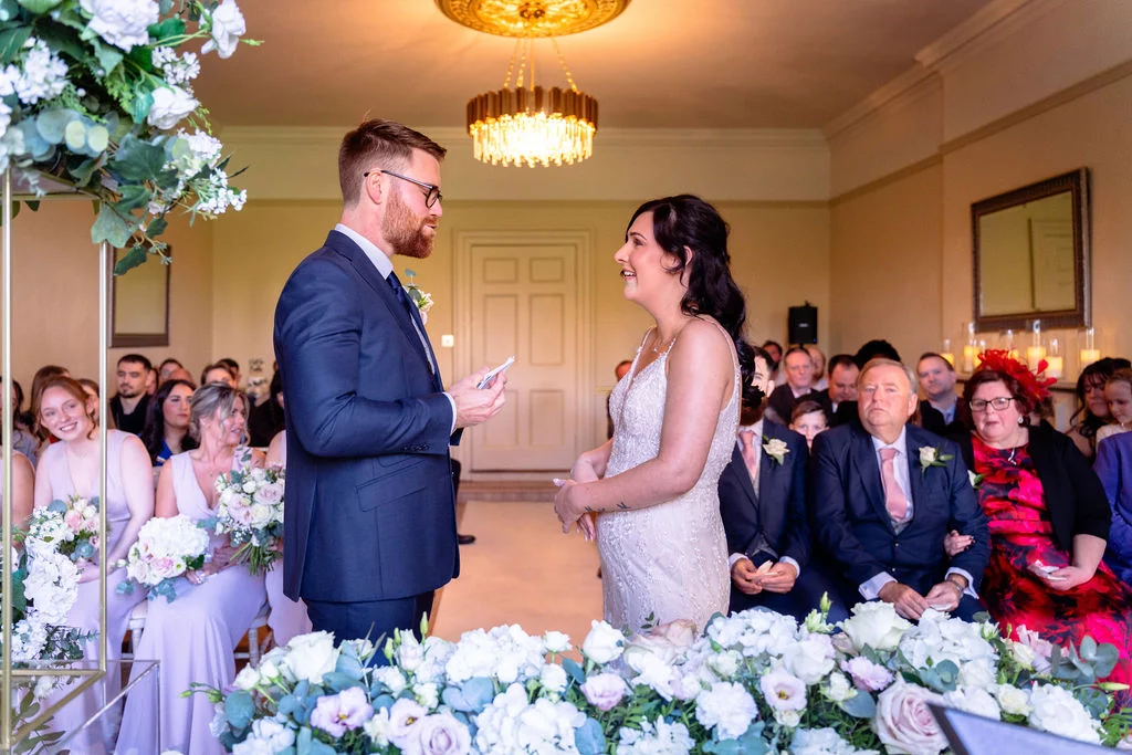 A groom in a dark suit reads his vows from a small card to his smiling bride during an indoor civil ceremony surrounded by seated guests and floral arrangements.