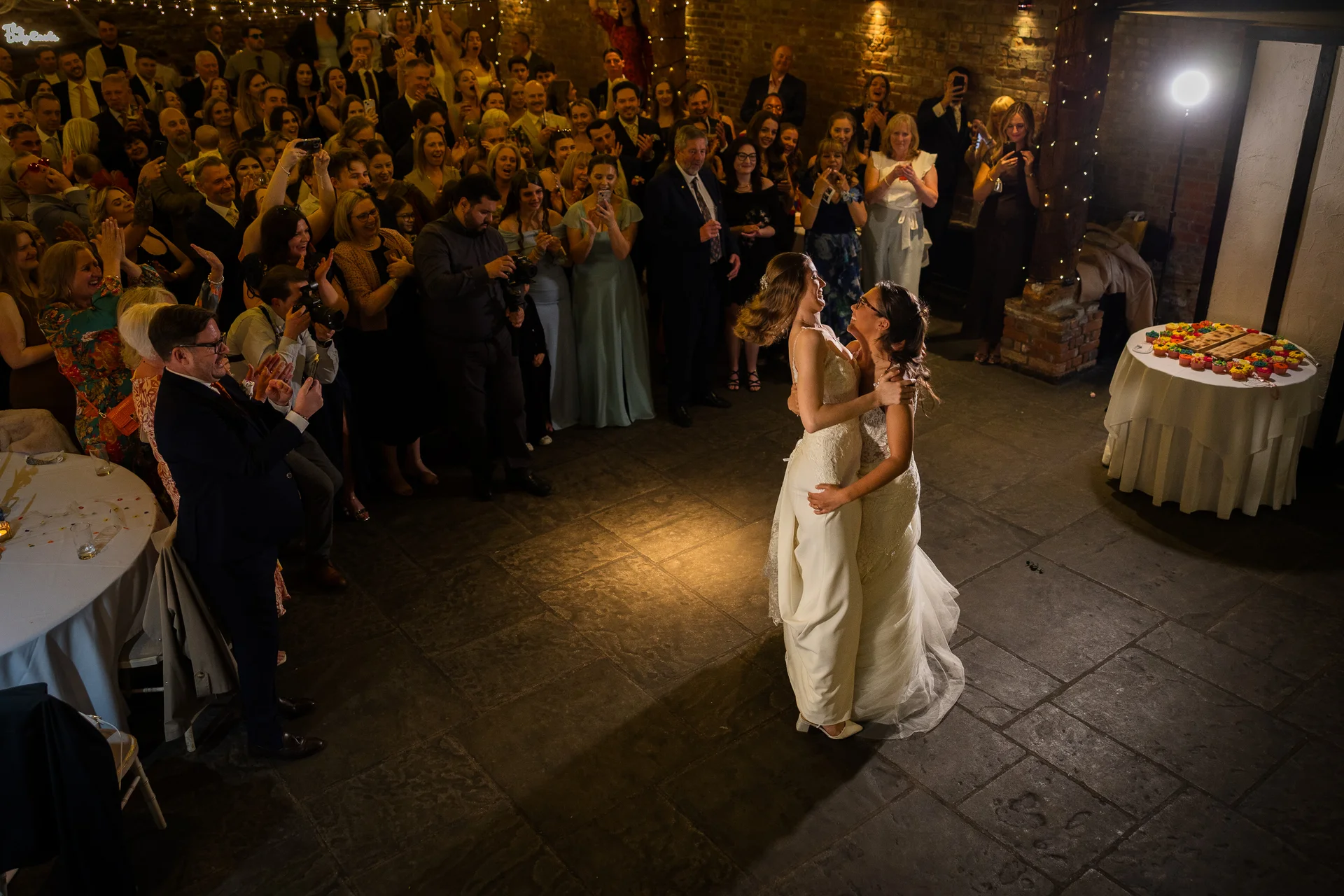 Two brides share a joyful first dance together on a stone floor while cheering wedding guests look on from the surrounding rustic venue.
