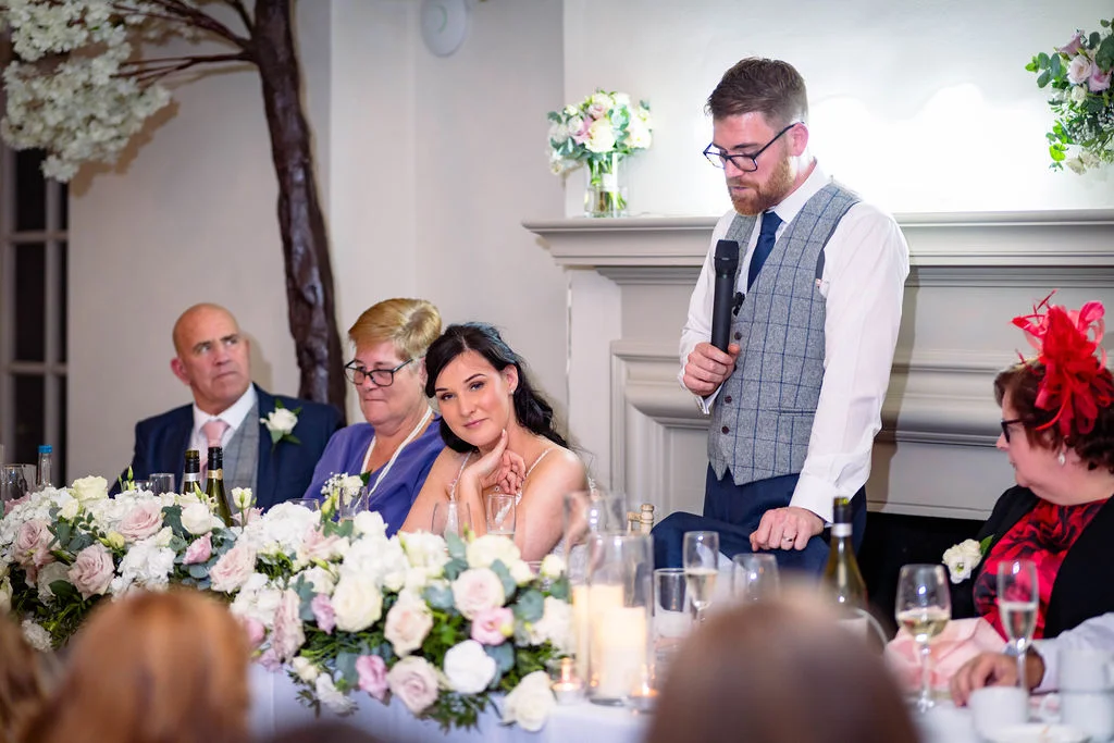 A groom delivers a speech at the top table during a wedding reception, while the bride and family look on amidst floral centrepieces and candles.