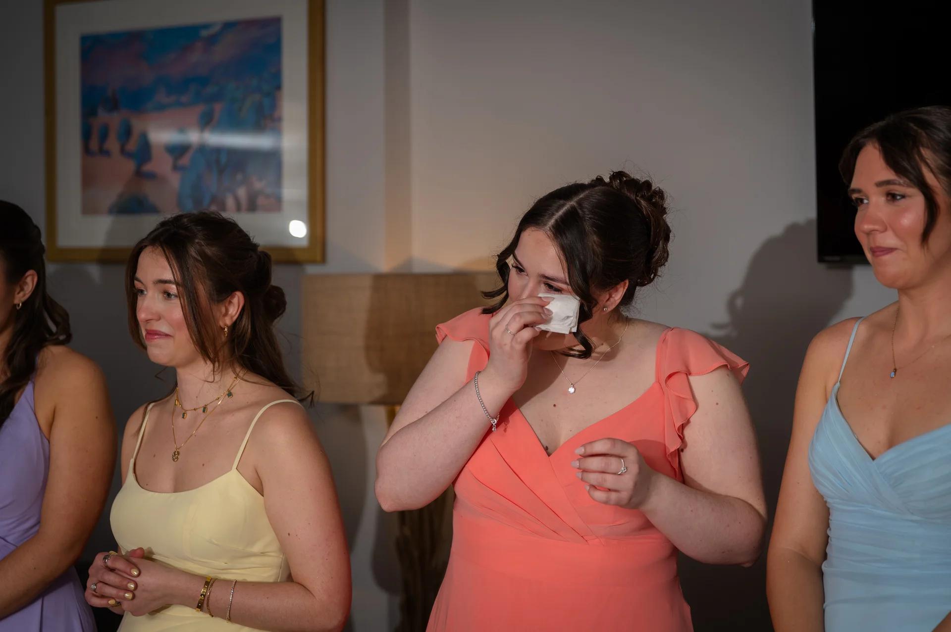 An emotional bridesmaid wiping a tear with a tissue while surrounded by her fellow bridesmaids in pastel-coloured dresses.