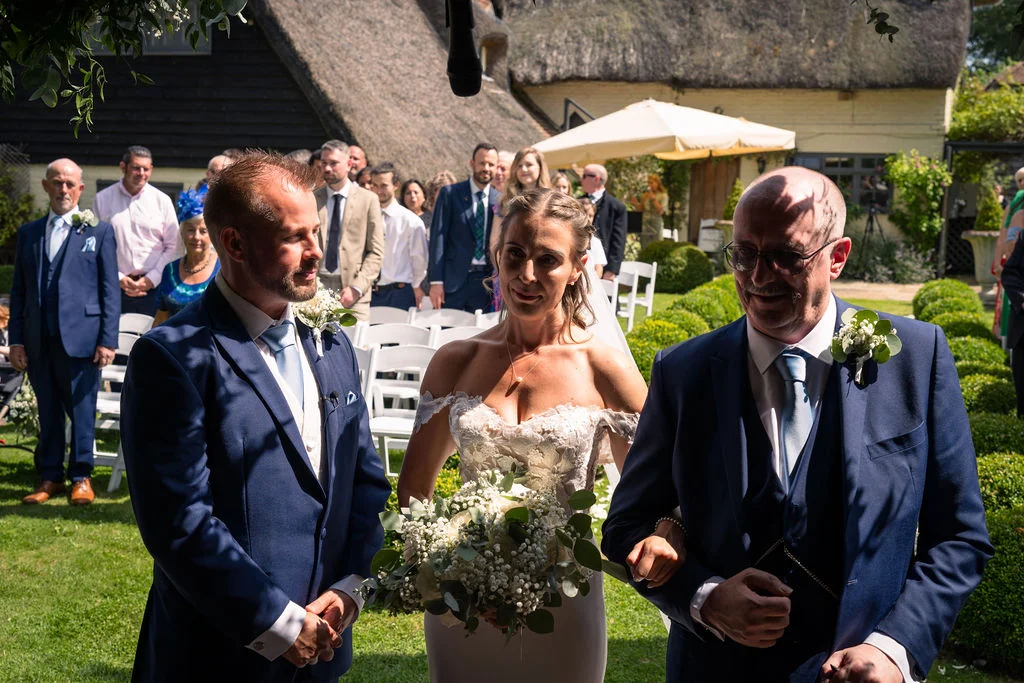 A smiling bride holding a white floral bouquet walks arm in arm with her father alongside the groom during an outdoor sunny wedding ceremony.