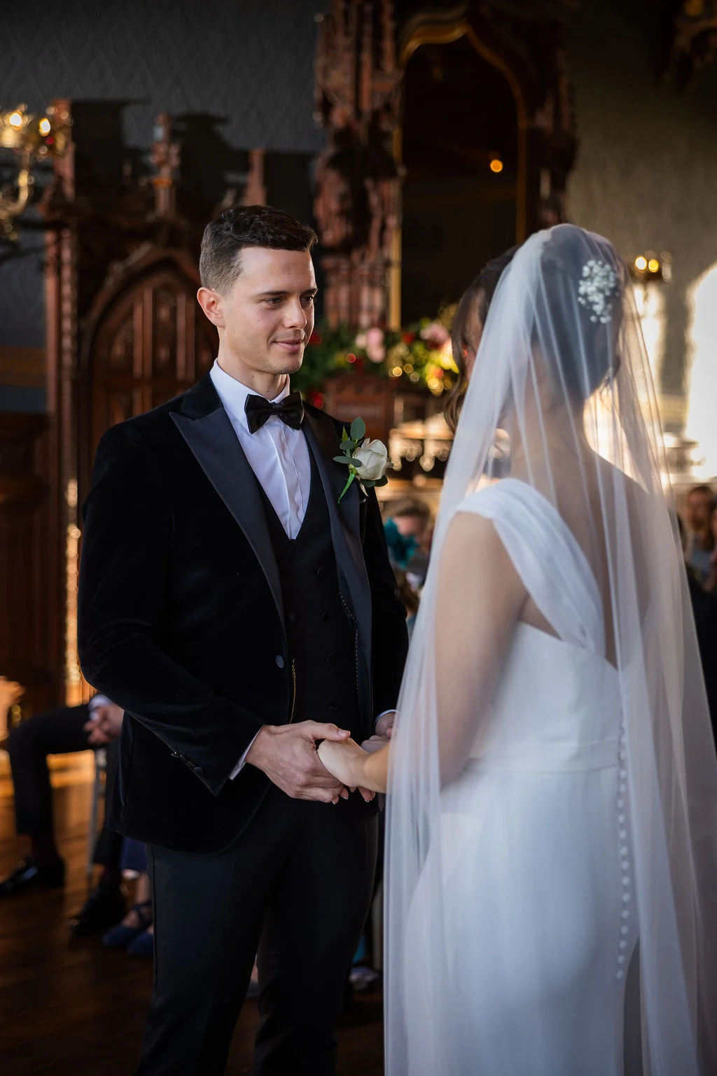 The groom looks lovingly at his bride while holding her hands during their ceremony inside a historic venue.