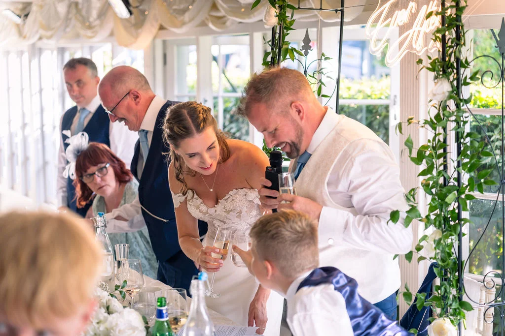 A groom delivers a speech while holding a microphone at the top table, making the smiling bride and guests laugh during the wedding reception.