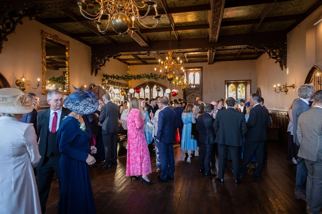A bustling drinks reception fills a historic panelled hall with guests mingling under ornate chandeliers.