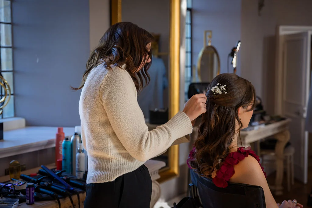 A professional hair stylist meticulously places delicate baby's breath flowers into a bride's half-up wavy hairstyle during wedding morning preparations.