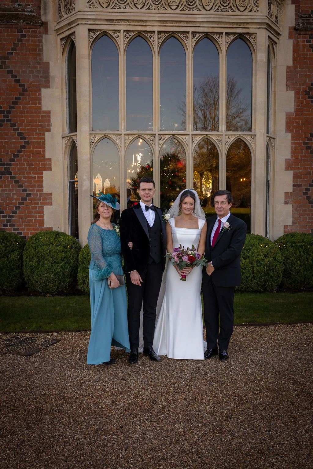 A newlywed couple poses outside a historic manor house with their parents on a gravel pathway in front of large arched windows.