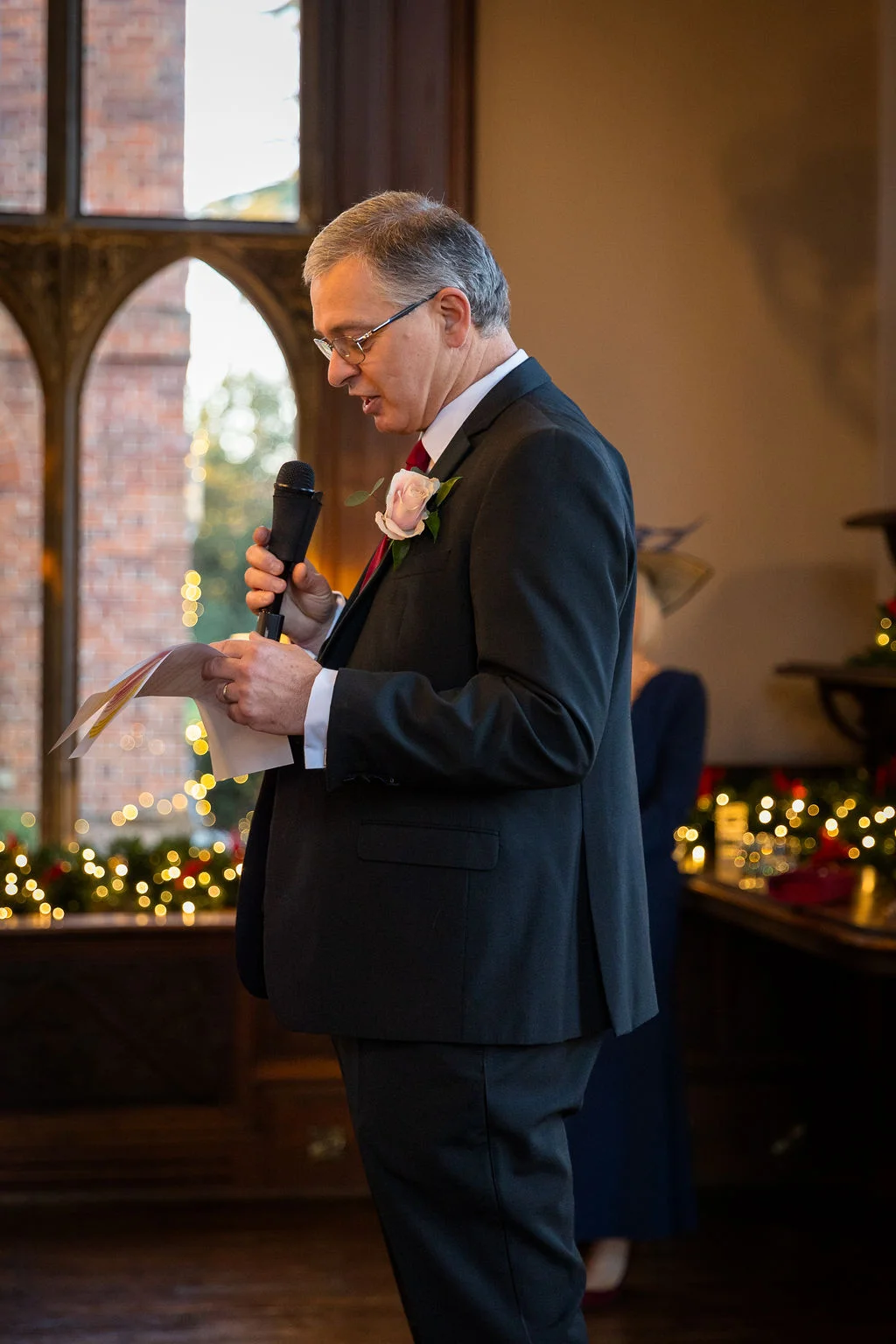 A groom delivers a heartfelt speech during the wedding reception, holding a microphone and notes in front of a softly lit window.
