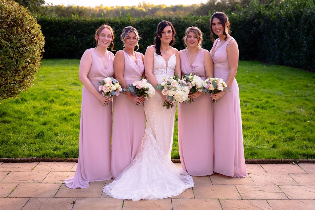 A bride in a lace fit-and-flare gown stands outdoors on a stone patio with four bridesmaids in matching floor-length pink dresses, all holding hand-tied floral bouquets during golden hour.