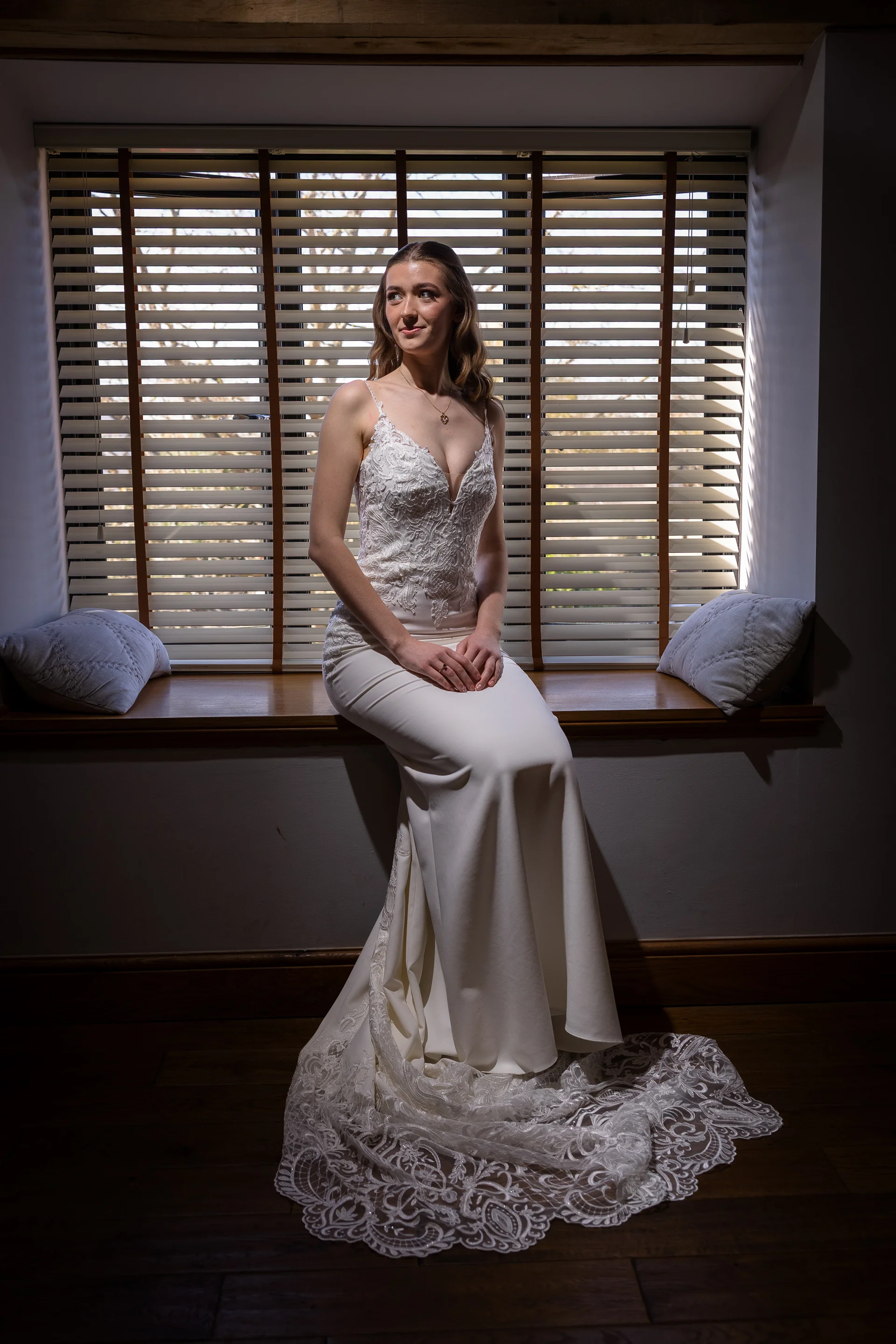 A stunning bride sits gracefully on a wooden window seat bathed in natural backlight, wearing a fitted crepe gown with a plunging lace bodice and delicate spaghetti straps.