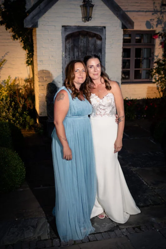 Two brides pose together outdoors in the evening outside a white brick building, one wearing a strapless lace-topped wedding gown and the other in a flowing dusty blue dress.