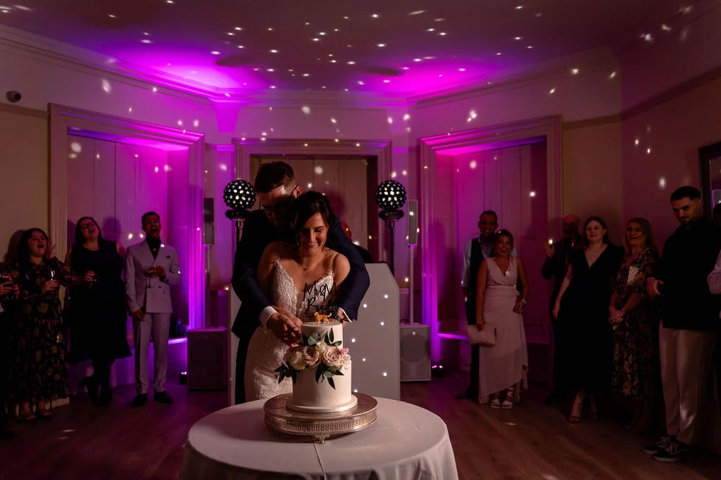 A bride and groom cut their white tiered wedding cake together while surrounded by cheering guests under dramatic pink disco lighting.