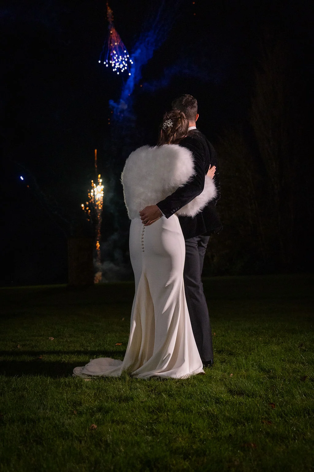 A newlywed couple embraces outdoors at night on a lawn, watching a colourful fireworks display light up the dark sky.