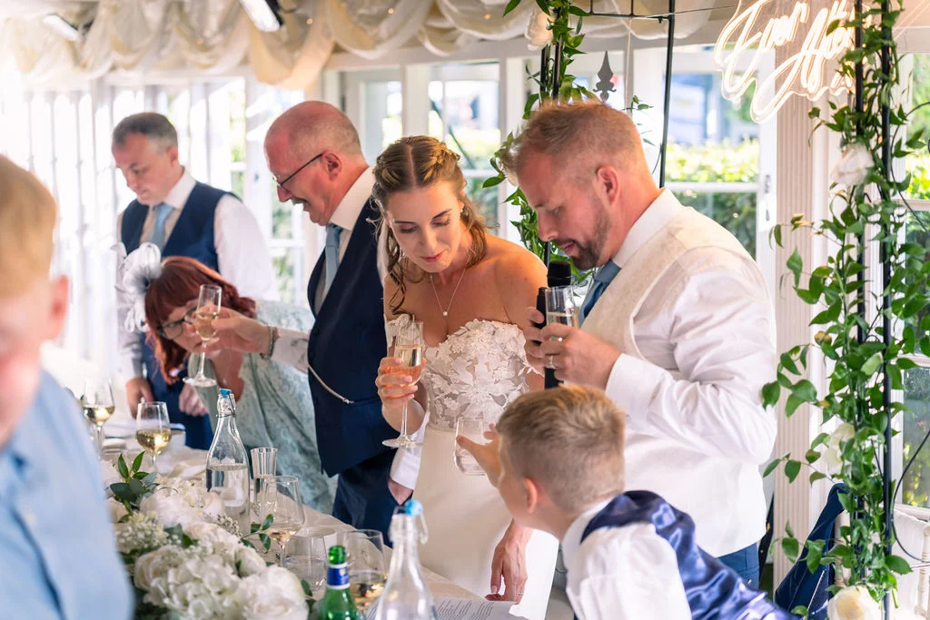 The groom delivers a speech at the top table while the bride and guests hold champagne flutes and share a laugh during the wedding reception.